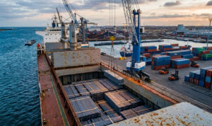 Aerial view of shipping steel billets in a bulk carrier hold at an industrial port with container terminals in the background, illustrating global steel logistics.
